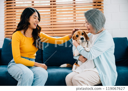On the sofa, a happy woman and her mother embrace their Beagle dog, creating an affectionate family portrait filled with togetherness and smiles. Their bond reflects the strength of family friendships On the sofa, a happy woman and her mother embrace their Beagle dog, creating an affectionate family portrait filled with togetherness and smiles. Their bond reflects the strength of family friendships 119067627