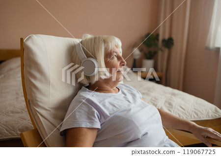 Elderly 60s woman in headphones lying on chair and listening to music with closed eyes while resting in cozy living room at home Elderly 60s woman in headphones lying on chair and listening to music with closed eyes while resting in cozy living room at home 119067725
