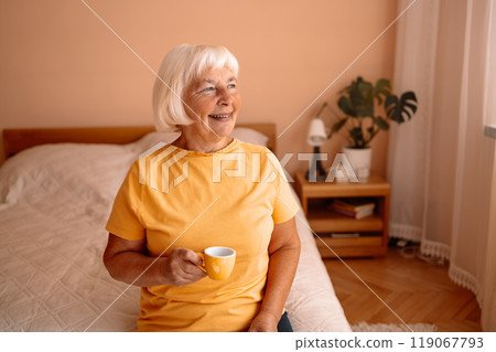 Caucasian blonde senior woman sitting on a white bed with a cup in her hand. Female relaxing and drinking cup of hot coffee or tea in the bedroom. 119067793