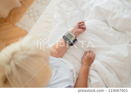 Close up of woman checks her blood pressure, a tonometer is worn on her wrist in a bed. Medical device for measuring blood pressure and heart rate used at hand wrist Close up of woman checks her blood pressure, a tonometer is worn on her wrist in a bed. Medical device for measuring blood pressure and heart rate used at hand wrist 119067801