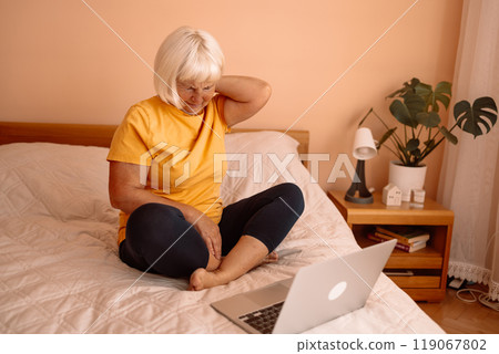 Overtired business senior 60s woman working with laptop on a bed, in morning. Female sitting on bed and touching her lower back and neck, experiencing body aches, wearing pajamas. 119067802