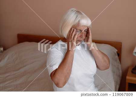 Stressed frustrated middle aged lady suffering from headache massaging temples sits on a bed. Tired sick mature old woman feeling unwell, touching head. Migraine pain, fatigue, stress concept 119067809