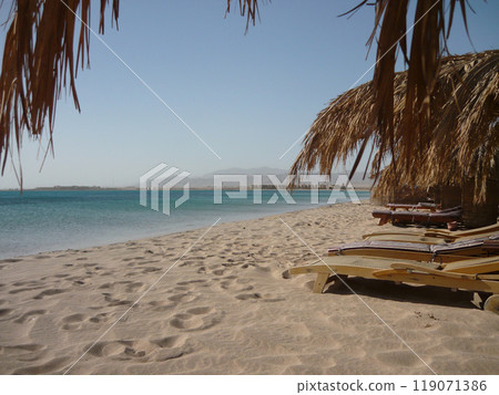 An empty sandy beach with palm leaf sunshade and wooden beds, blue sea view without waves An empty sandy beach with palm leaf sunshade and wooden beds, blue sea view without waves 119071386