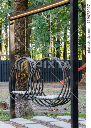 Modern hanging round swings in a cozy garden setting, surrounded by lush green trees and sunlight 119072067