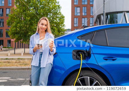 Cheerful woman with a coffee in hand checks her EV battery level and completes the payment using a charging app on the street Cheerful woman with a coffee in hand checks her EV battery level and completes the payment using a charging app on the street 119072659