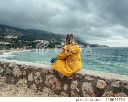 A woman in a yellow windbreaker looks out from a cliff at the sea A woman in a yellow windbreaker looks out from a cliff at the sea 119075734