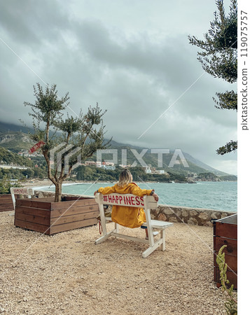 A woman in a yellow raincoat sits on a bench 119075757
