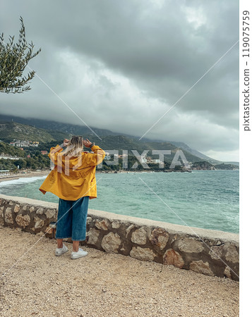 A woman in a yellow windbreaker looks out from a cliff at the sea 119075759