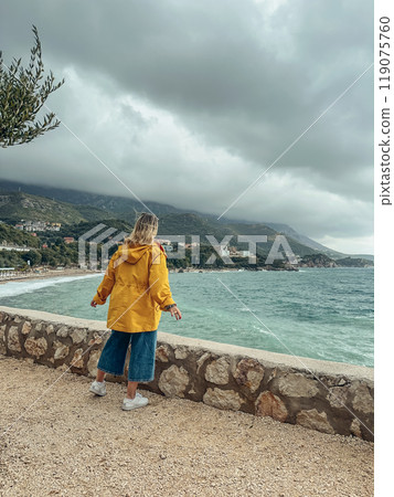 A woman in a yellow windbreaker looks out from a cliff at the sea A woman in a yellow windbreaker looks out from a cliff at the sea 119075760