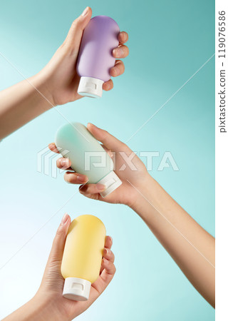 Three hands are shown in this skin care product advertising gently gripping and raising three unbranded lotion bottles in shades of blue, yellow, and purple on a crisp blue backdrop. 119076586