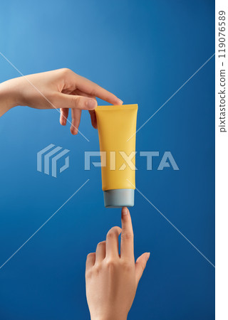 A unlabeled yellow tube of cream is held upright by a model in a blue sky, one hand holding the base and the other touching the cap. A minimalist photo composition for a skincare product advertisement 119076589