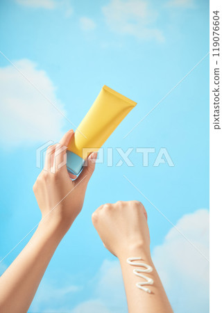 An unbranded yellow tube with a blue cream cap is held by a model, who is wearing a smear of cream on wrist, suitable for promoting a skincare product. There are clouds and a blue sky in the backdrop. An unbranded yellow tube with a blue cream cap is held by a model, who is wearing a smear of cream on wrist, suitable for promoting a skincare product. There are clouds and a blue sky in the backdrop. 119076604