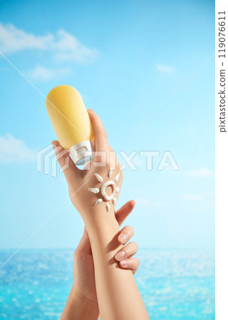 A female model hands are gently clasped together and holding a tube of unlabeled lemon yellow cream. On the back of her hand is a sun-shaped cream mark, suitable for advertising images for sunscreen. 119076611