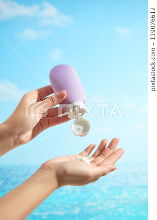 The backdrop of a sunny blue sky and sea features a female model hands squeezing an unbranded purple bottle of cream into a hand, which is perfect for a sunscreen advertisement image. The backdrop of a sunny blue sky and sea features a female model hands squeezing an unbranded purple bottle of cream into a hand, which is perfect for a sunscreen advertisement image. 119076612