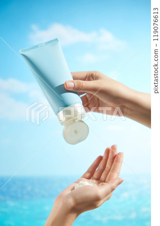 An image of a female hand pressing an unbranded blue tube of cream into a white cream against the backdrop of a serene, sunny blue sky and sea is ideal for advertising for sunscreen. 119076613