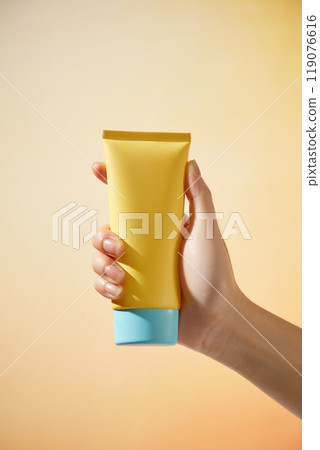 Minimalist composition image on a bright orange background resembling the sun with the model hand holding an unlabeled yellow tube with a blue cap, suitable for sunscreen advertisements. Minimalist composition image on a bright orange background resembling the sun with the model hand holding an unlabeled yellow tube with a blue cap, suitable for sunscreen advertisements. 119076616