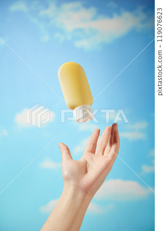 A close-up image of a yellow squeeze bottle with a white cap floating in the air. A female hand is reaching up to grab the bottle. The background is a bright blue sky with white clouds. A close-up image of a yellow squeeze bottle with a white cap floating in the air. A female hand is reaching up to grab the bottle. The background is a bright blue sky with white clouds. 119076623