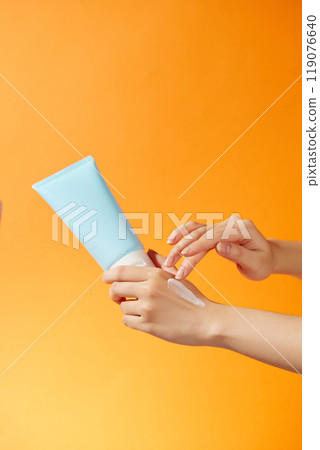 The model gently applies cream to the back of her hand from a mockup blue tube held by the model against a bright orange background like the sun, perfect for a sunscreen advertisement. The model gently applies cream to the back of her hand from a mockup blue tube held by the model against a bright orange background like the sun, perfect for a sunscreen advertisement. 119076640