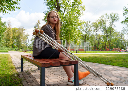 Melancholy teen in casual dress sits dejectedly on park bench, orange cast on leg, crutches by her side - her pensive gaze reflects inner turmoil Melancholy teen in casual dress sits dejectedly on park bench, orange cast on leg, crutches by her side - her pensive gaze reflects inner turmoil 119078386