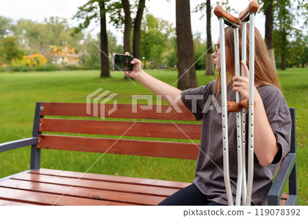 Teenage girl with a leg cast seated on a park bench, capturing a bittersweet selfie as a memento of her injury and the time lost during the recovery process 119078392
