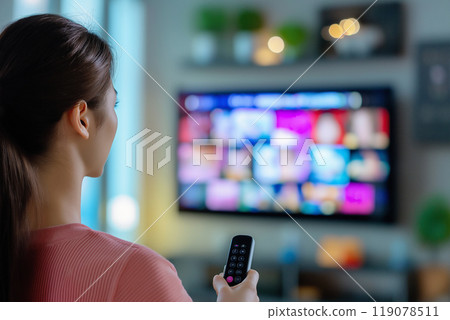 Rear view of an Asian woman holding a TV remote control and choosing a video to watch 119078511