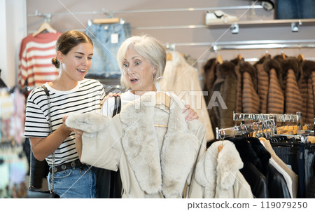 Girl consults with senior female friend about quality fur coat during shopping in clothes store Girl consults with senior female friend about quality fur coat during shopping in clothes store 119079250