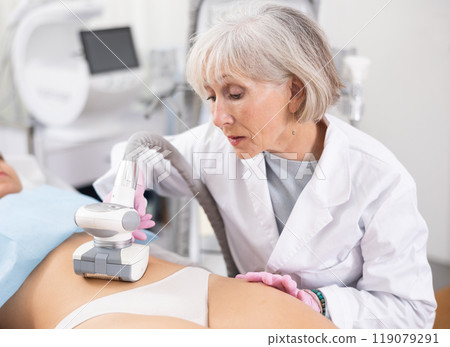 Elderly female doctor doing Rf lifting procedure on belly of young woman in beauty salon. Treatment of flabby skin Elderly female doctor doing Rf lifting procedure on belly of young woman in beauty salon. Treatment of flabby skin 119079291