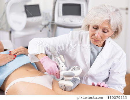 Old woman specialist doing LPG massage of hips of young woman Old woman specialist doing LPG massage of hips of young woman 119079313