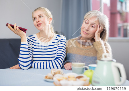 Adult daughter and elderly mother talking on phone and texting at home 119079339