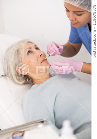 Young woman cosmetologist in uniform making injections for lip augmentation and anti-wrinkle of elderly female in esthetic clinic indoors 119079379