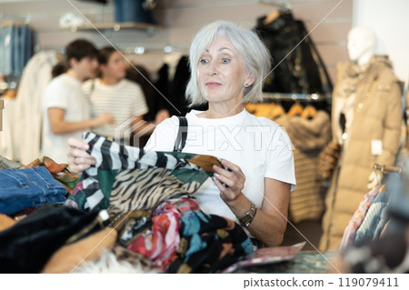 Old woman choosing second hand clothes in large clothing shop 119079411