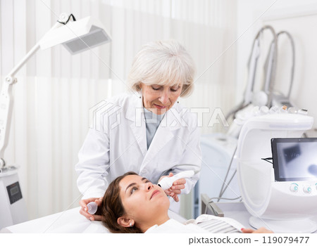 Professional mature female cosmetologist doing facial skin ultrasound cavitation for flawless woman face laying in beauty salon 119079477