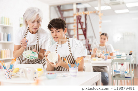 Elderly woman assisting adult son painting ceramic mugs in family workshop 119079491
