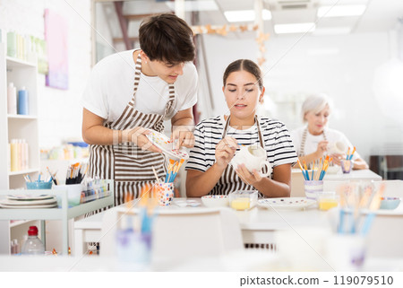 Guy assisting young girl painting ceramic mugs in pottery class 119079510