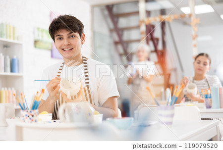 Happy young guy painting on ceramic cup during pottery class 119079520