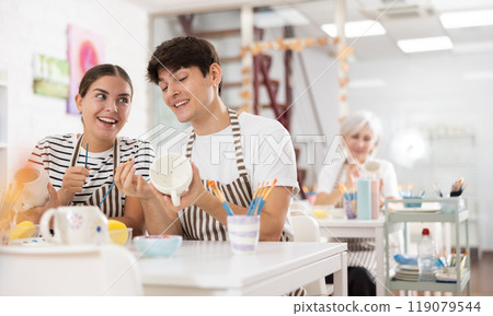 Young couple joyfully talking and painting ceramic mugs in pottery workshop Young couple joyfully talking and painting ceramic mugs in pottery workshop 119079544