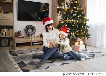 Mother and daughter use phone in front of Christmas tree at home 119080718