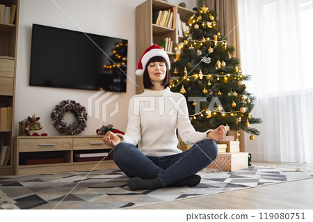 Young woman meditating by Christmas tree in cozy home setting 119080751