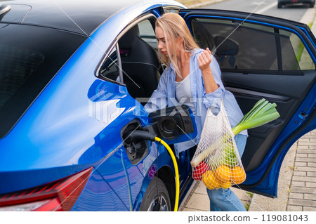 Woman plugs in her electric vehicle to charge while holding a bag filled with fresh groceries. Woman plugs in her electric vehicle to charge while holding a bag filled with fresh groceries. 119081043
