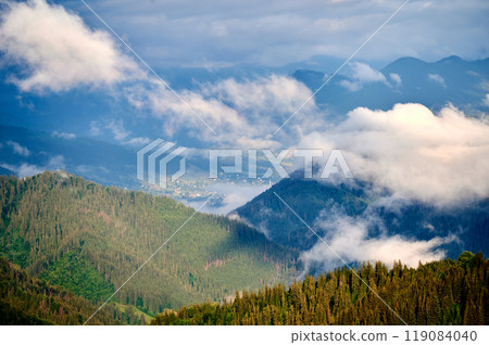 Picturesque scene of mountain valley blanketed in mist, with patches of green pastures and scattered houses. Clouds drift gently over forested hills, creating serene and tranquil atmosphere. 119084040