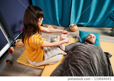 Masseuse making thai yoga massage. Woman therapist doing traditional massage treatment, stretching male patient's body. Client lying on mat, enjoying hand massage in calm atmosphere. 119084043