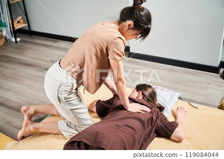 Masseuse making thai yoga massage. Woman therapist doing traditional massage treatment for female patient indoors. Client lying on mat, meditating, enjoying hand massage in calm atmosphere. 119084044