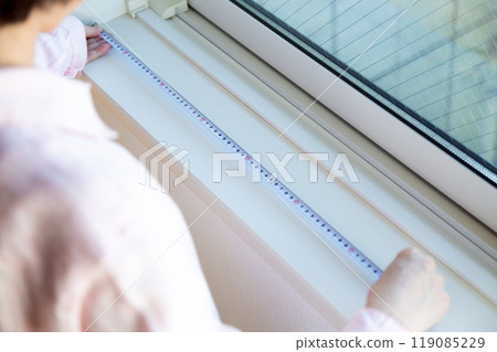 A middle-aged woman measuring the size of curtains at a window in an apartment A middle-aged woman measuring the size of curtains at a window in an apartment 119085229