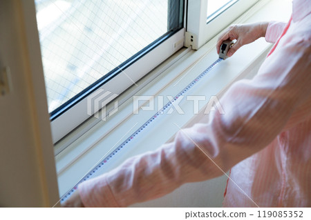 A middle-aged woman measuring the size of curtains at a window in an apartment A middle-aged woman measuring the size of curtains at a window in an apartment 119085352