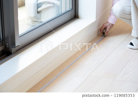 A middle-aged woman measuring the size of curtains at a window in an apartment 119085353