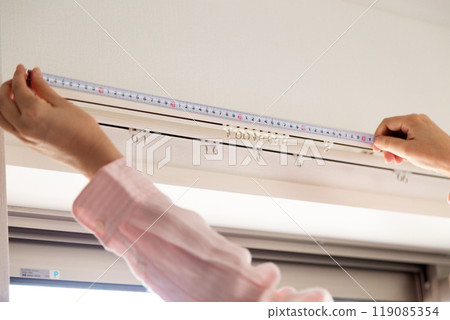 A middle-aged woman measuring the size of curtains at a window in an apartment A middle-aged woman measuring the size of curtains at a window in an apartment 119085354
