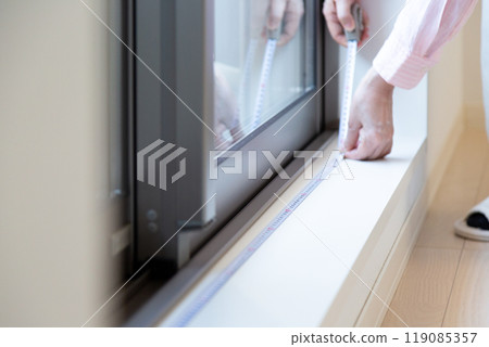 A middle-aged woman measuring the size of curtains at a window in an apartment A middle-aged woman measuring the size of curtains at a window in an apartment 119085357