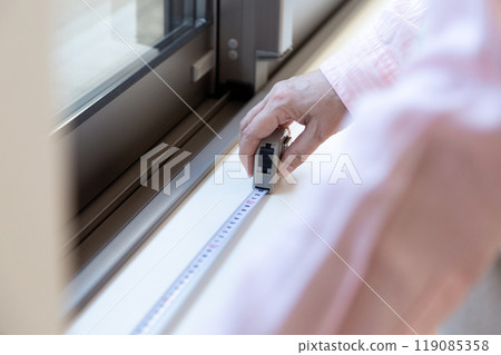A middle-aged woman measuring the size of curtains at a window in an apartment A middle-aged woman measuring the size of curtains at a window in an apartment 119085358