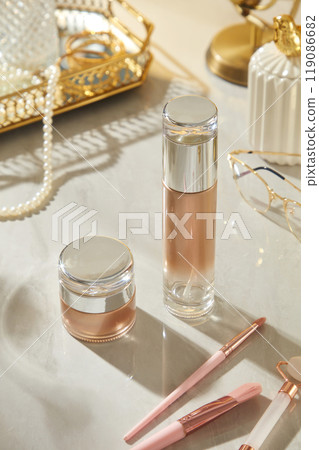Two skincare products, a bottle and a jar, are placed on a white surface with a pink brush and a jade roller. The image exudes luxury, ideal for showcasing beauty products and branding elements. 119086682