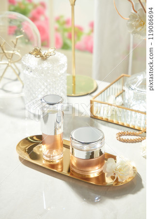 A top-down photo depicts two unlabeled lotion bottles on a brass tray with white flowers, surrounded by glass boxes, jewelry, and gold-colored ornaments, set against a luxurious backdrop of white silk 119086694